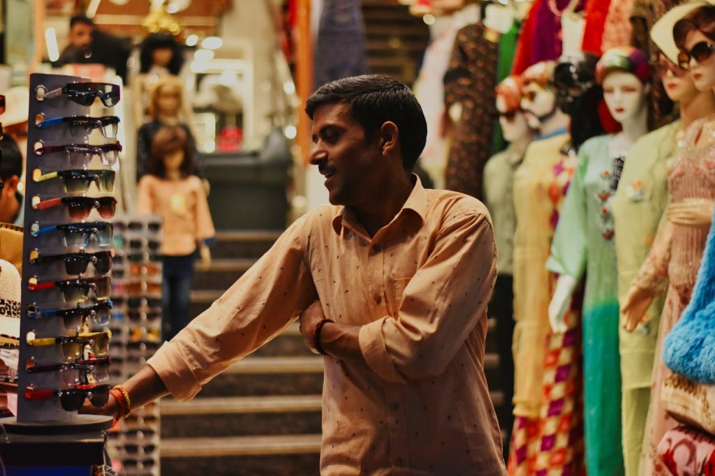 Man smiling in a vibrant clothes store in Katra, standing by a sunglasses display.